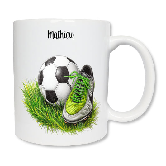 Mug blanc en céramique de 325 ml avec le dessin d'un ballon de foot et une chaussure de foot posés sur de l'herbe, et personnalisé avec un prénom. Cadeau pour footballeur ou coach.