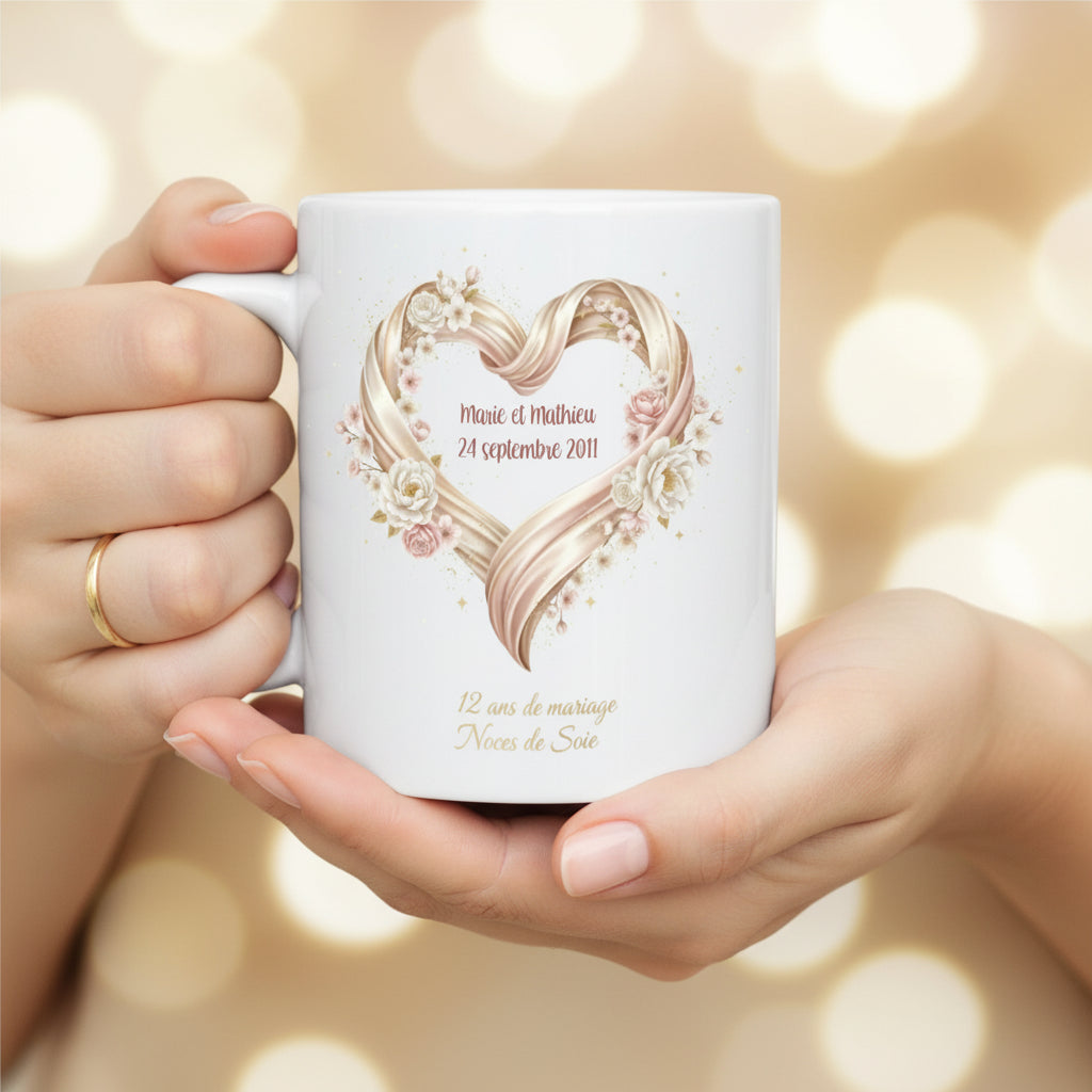 Mug personnalisé 12 ans de mariage Noces de Soie, tenu à deux mains sur fond bokeh doré, illustration cœur en ruban de soie doré et roses blanches
